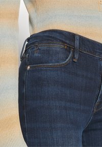 Jeans de mezclilla azul oscuro con corte slim y diseño de cinco bolsillos, con costuras doradas y una cinturilla recta. Textura suave visible.