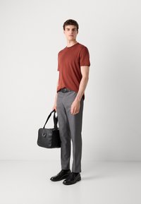 Tricou scurt de culoare ruginită, pantaloni gri, curea neagră și pantofi din piele lucioasă negri, ținând o geantă neagră cu curea.