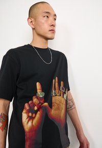 Černé tričko s krátkým rukávem s grafikou dvou rukou s několika prsteny. Materiál vypadá měkký a má pohodlný střih.