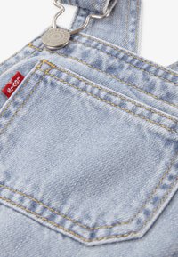 Φωτεινά μπλε τζιν φορμάκια με ασημένια μεταλλικά στοιχεία, πορτοκαλί ραφές και κόκκινη ετικέτα μάρκας στην μπροστινή τσέπη, με κλασικό σχέδιο τσέπης.