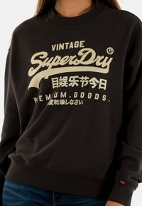 Felpa nera con collo e polsini a costine. Presenta il logo dorato stampato "SuperDry" e testo giapponese. Materiale in misto cotone, vestibilità rilassata.
