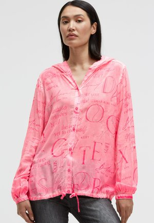 Frau mit langen dunklen Haaren, die ein leuchtend pinkes, kapuzenbesetztes Hemd mit Knöpfen und motivierendem Text trägt, dazu dunkle Jeans, stehend vor einem schlichten Hintergrund.