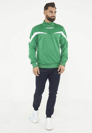 Uomo con la barba che indossa una giacca sportiva verde LEGEA, pantaloni neri e sneakers bianche, in piedi su uno sfondo bianco uniforme.