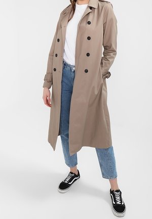 Femme portant un trench-coat beige à double boutonnage, un t-shirt blanc, un jean bleu raccourci et des baskets noires avec des lacets blancs, debout avec une main dans la poche.