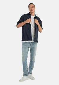 Camisa de manga corta navy sobre una camiseta blanca, combinada con unos vaqueros de jeans claros y zapatillas blancas. Presenta dos bolsillos en el pecho y detalles de botones.