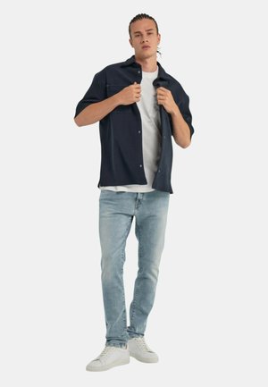 Camisa de manga corta navy sobre una camiseta blanca, combinada con unos vaqueros de jeans claros y zapatillas blancas. Presenta dos bolsillos en el pecho y detalles de botones.