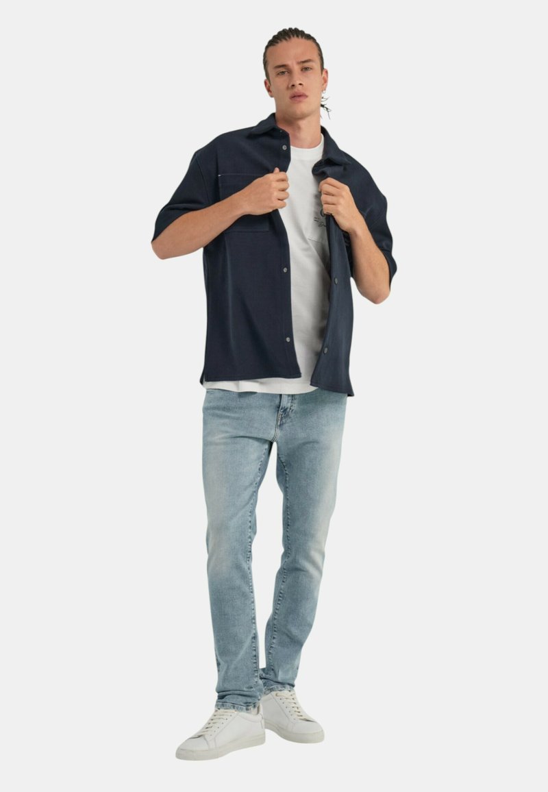 Camisa de manga corta navy sobre una camiseta blanca, combinada con unos vaqueros de jeans claros y zapatillas blancas. Presenta dos bolsillos en el pecho y detalles de botones.