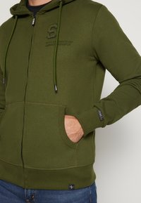 Olivgrüner Reißverschluss-Hoodie aus weichem Stoff mit einer vorderen Kängurutasche und gerippten Bündchen. Mit einem Logo auf der Brust und einer Kapuze mit Kordelzug.
