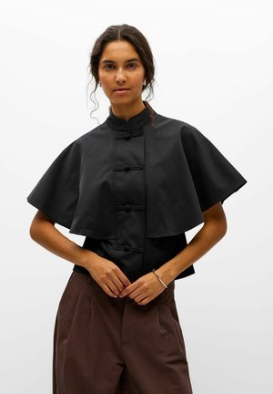 Une femme porte une veste noire de style cape avec des brandebourgs et un pantalon marron sur un fond blanc uni.