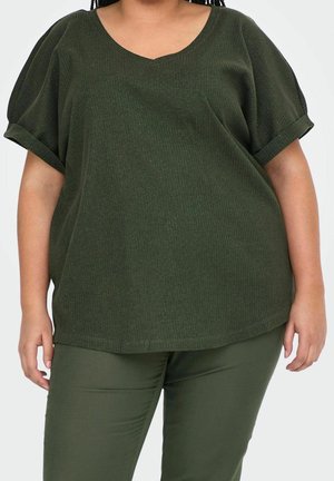 Personne portant un haut côtelé vert foncé à manches courtes et col en V, avec un pantalon ajusté assorti sur fond blanc.