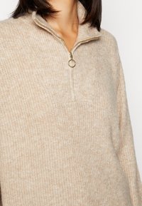 Frau trägt einen beigen Strickpullover mit einem goldenen Reißverschluss und rundem Griff am Hals, zeigt den Oberkörper und einen Teil der Haare.