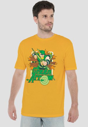 Mann trägt gelbes T-Shirt mit buntem Green Arrow-Grafik, hält Bogen und Pfeil, steht vor hellem einfarbigem Hintergrund.