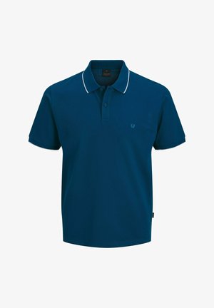 Giovane persona in piedi, indossa una polo verde scuro, pantaloncini di jeans grigi risvoltati, calzini bianchi e scarpe da ginnastica bianche, su uno sfondo semplice.
