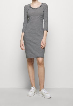 Kobieta stoi w czarno-białej, paskowanej sukience do kolan z rękawami 3/4 oraz białych sneakersach z złotymi akcentami.