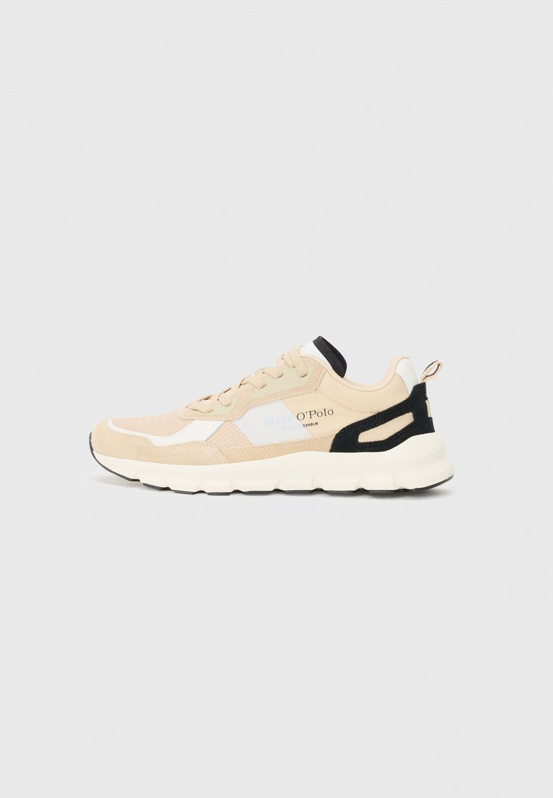 Zapatillas deportivas beige con materiales de malla y gamuza, acentos negros y una suela blanca gruesa. Presenta una marca en el lateral y una pestaña de sujeción.