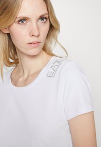 Camiseta de algodón blanca con cuello redondo y mangas cortas, que presenta el logotipo en gris "EA7 Emporio Armani" en el hombro. Textura suave.