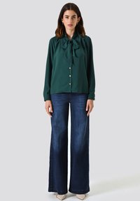 Blusa verde con dettaglio del fiocco, maniche lunghe e bottoni argentati, abbinata a jeans in denim scuri a gamba larga e tacchi a punta di colore chiaro.