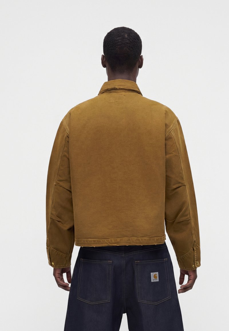 Man wearing a brown jacket and dark blue denim pants, shown from the back against a plain background.