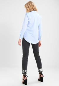 Camisa de rayas azul claro con un dobladillo curvado, combinada con pantalones gris oscuro que tienen texto blanco en los puños y tacones negros.