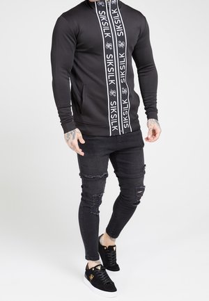 Mężczyzna w czarnej bluzie SikSilk na zamek z paskami z logo, podartych czarnych obcisłych dżinsach i czarnych sneakersach ze złotymi detalami.