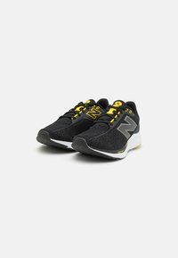 Un par de zapatillas de correr negras con acentos amarillos y logotipos grises, suelas blancas acolchadas y tela de malla texturizada, colocadas sobre un fondo blanco liso.