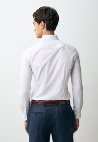 Maglietta bianca a maniche lunghe con colletto classico e polsini con bottoni, abbinata a pantaloni scuri e cintura in pelle marrone.