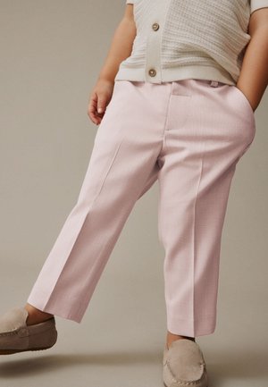 Barn iført pink tilpassede bukser, beige strikket knap-top og beige slip-on sko, stående med den ene hånd i lommen.