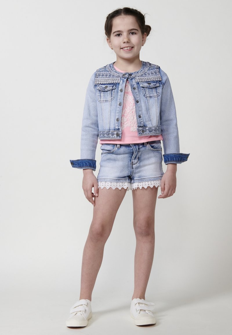 Denim jakna z vzorom na yoku, roza majica, denim kratke hlače z guipure čipko in bele superge. Svetle barve in sproščen dizajn.