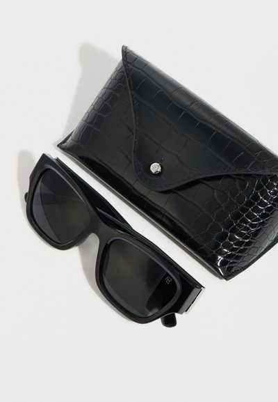 Μεγαλύτερα από το κανονικό μαύρα τετράγωνα γυαλιά ηλίου με σκουρόχρωμους φακούς, τοποθετημένα δίπλα σε μαύρη δερμάτινη θήκη με σχέδιο κροκοδείλου και ασημένιο κουμπί κλεισίματος.
