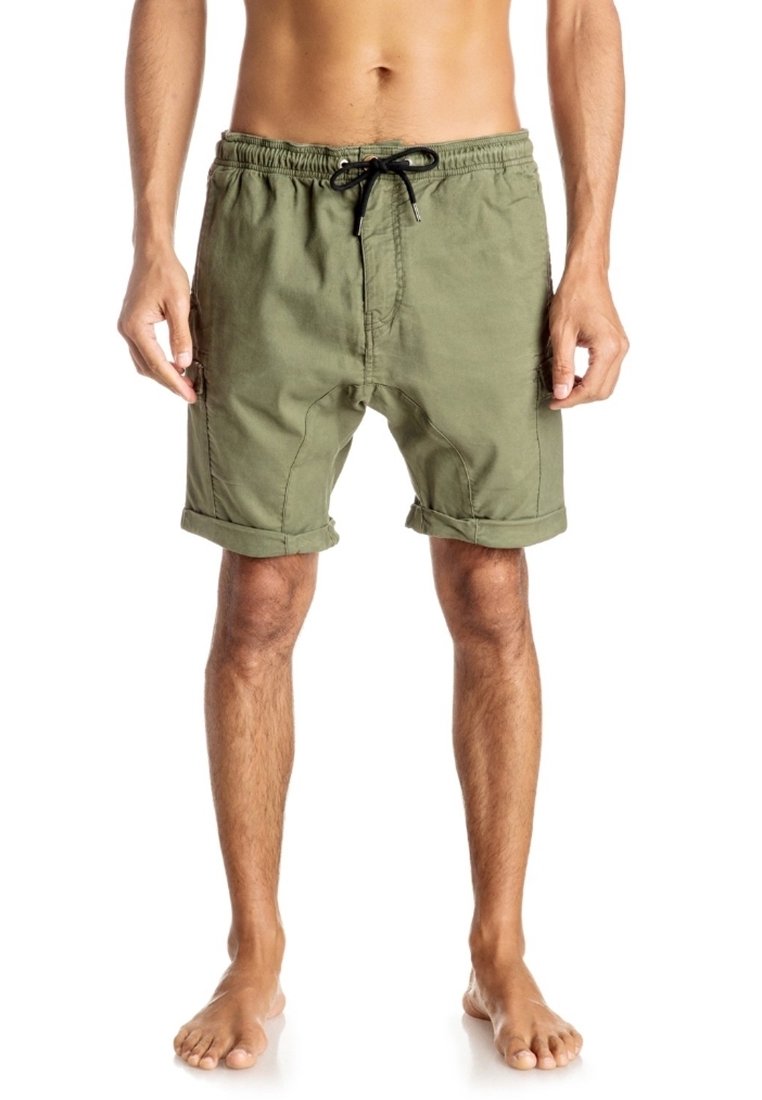 Homme pieds nus portant un short vert olive à cordon de serrage avec revers roulés, debout devant un fond blanc, les mains détendues le long du corps.