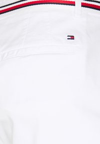 Tessuto bianco con una superficie liscia. Presenta una cintura a strisce rossa, blu e bianca e un piccolo logo nell'angolo. Design semplice, stile casual.