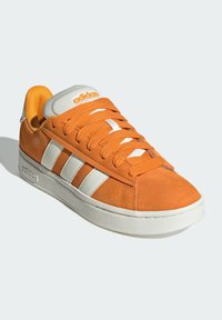 Zapatilla Adidas de ante naranja con rayas blancas, cordones naranjas, suela blanca y logo en la lengüeta y el costado, exhibida sobre un fondo blanco.
