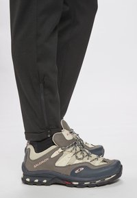 Person som bär svarta byxor med ankelsidöppningar och grå-beiga Salomon vandringsskor med svarta och röda sulor på en vit bakgrund.