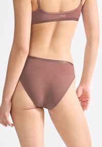 Vrouw in een mauve naadloze bh en bijpassende hoge onderbroek gezien vanuit de achterkant tegen een plain witte achtergrond.