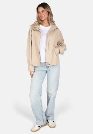 Veste à capuche beige avec fermeture éclair et pression, portée sur un t-shirt blanc. Associée à un jean bleu clair et des baskets blanches.