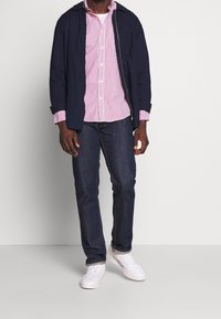 Veste marine portée sur un t-shirt à rayures rouges et blanches, associée à un jean foncé et des baskets blanches. Design simple avec des couleurs contrastées.