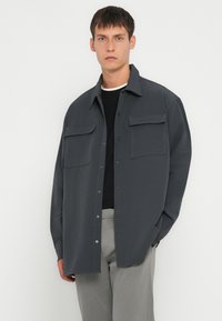 Graues Button-up-Hemd mit zwei Brusttaschen, aus einem glatten Material gefertigt. Verfügt über einen Kragen und sichtbare Nähte. Kombiniert mit neutralen Hosen.