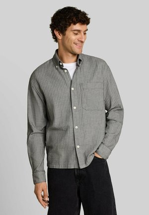 Uomo che sorride e guarda verso il basso indossando una camicia grigia a righe con bottoni sopra una t-shirt bianca con pantaloni neri contro uno sfondo semplice.