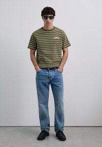 Camiseta a rayas color verde oliva, gafas de sol negras, jeans azules y zapatos negros. Diseño simple con mangas cortas y corte relajado.