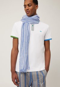 Vit polo t-shirt med gröna och blå randiga ärmar, grå knappslå och en blå- och vitrandig scarf runt halsen.