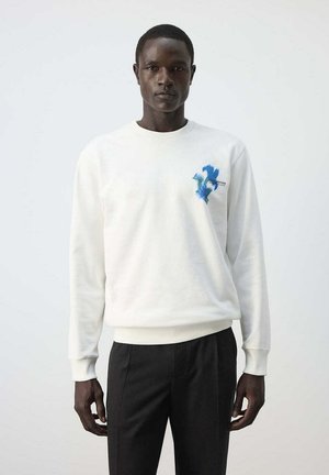 Homme portant un sweat-shirt blanc avec un petit motif floral bleu sur la poitrine et un pantalon noir, debout devant un fond clair uni.