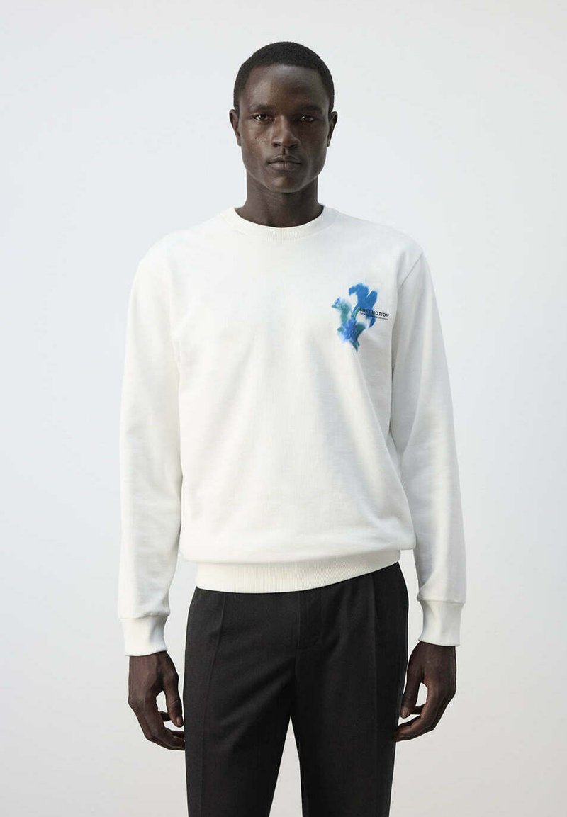 Homme portant un sweat-shirt blanc avec un petit motif floral bleu sur la poitrine et un pantalon noir, debout devant un fond clair uni.