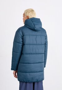 Πουκάμισο με επένδυση και κουκούλα σε σκούρο τιρκουάζ, με καπιτονέ σχέδιο, μακριά μανίκια και τσέπες. Διαθέτει ίσια γραμμή και λεία υφή.