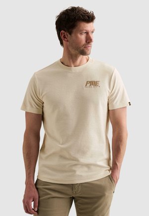 Hombre con camiseta beige de manga corta "PINE Original Aviator" y pantalones caqui, mirando hacia su izquierda con una mano en el bolsillo.