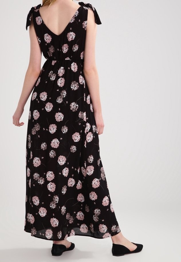 Femme portant une robe maxi noire à motif floral rose, bretelles nouées en nœud et chaussures plates noires, debout devant un fond uni.