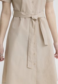 Beige jurk met knopen, korte mouwen en een stoffen ceintuur in de taille, gedragen door een persoon met ringen aan de vingers.