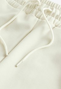 Pantaloni de trening bej deschis, confecționați din bumbac moale, cu o talie elastică, șnururi albe cu capete metalice argintii și o textură netedă.