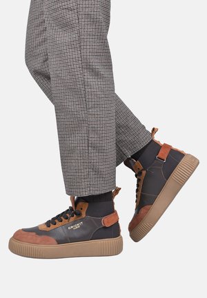 Personne portant des baskets montantes marron et noires avec des lacets noirs et des semelles beiges, associées à un pantalon gris à carreaux et des chaussettes noires.