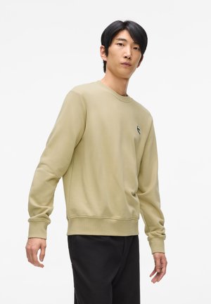 Felpa verde in cotone con colletto a girocollo, polsini e orlo ribattuti. Presenta un piccolo logo sul petto. Indossata con pantaloni scuri.