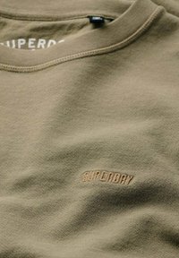 Sudadera verde oliva con una textura suave, cuello redondo y logotipo bordado "SUPERDRY" en color beige. Diseño simple sin patrones.
